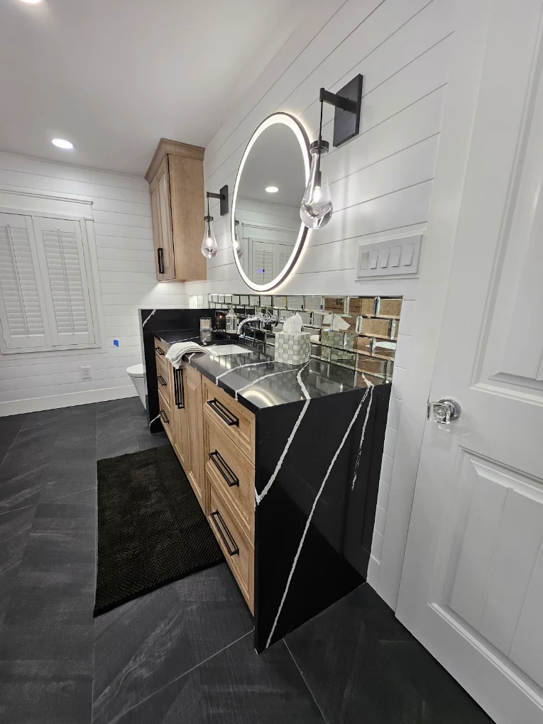 Wood bathroom vanity with a black waterfall countertop, round illuminated mirror, and white shiplap walls